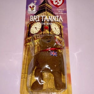 Puede incluir: Un oso de peluche marrón llamado "Britannia" en un embalaje de plástico transparente. El embalaje presenta una imagen del Big Ben y el texto "Collect All 4!". El oso tiene una cinta roja y una bandera de la Union Jack.