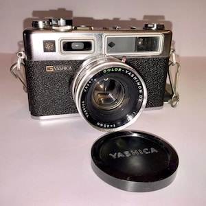 May include: A vintage Yashica camera with a silver and black body, a Color-Yashinon DX lens, and a lens cap. The camera has a strap and is set against a white background. The lens reads 1:1.7 f=45mm.