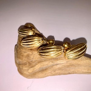 Puede incluir: Cuatro gemelos dorados con un diseño ovalado y acanalado. Los gemelos se exhiben sobre un trozo de madera flotante sobre un fondo rosa suave. La joyería tiene un acabado brillante.