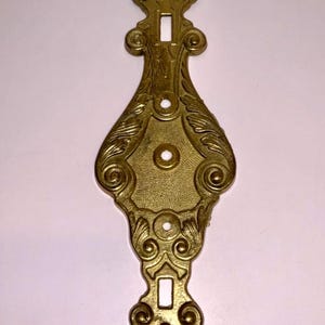 May include: Ornate brass-colored metal escutcheon plate with a symmetrical design. The plate features a central oval shape with decorative swirls and a keyhole at the top and bottom. The surface has a slightly aged appearance.
