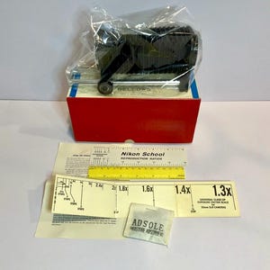 May include: A black Nikon bellows camera attachment, packaged in plastic, atop a red and blue box. Included are instruction sheets, a ruler, and a small packet labeled "ADSOLE MOISTURE ABSORBENT". The box is labeled "BELLOWS".