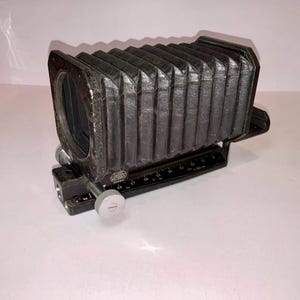May include: A black Leitz Wetzlar bellows attachment for a camera, featuring a pleated accordion-style design. The base includes a metal rail with printed measurement markings and a silver adjustment knob, with the brand name clearly visible.