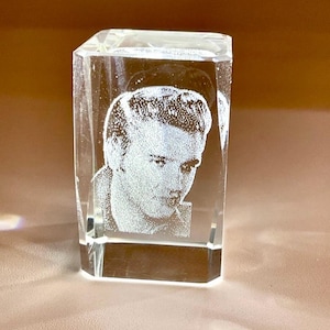 May include: A clear, rectangular crystal block with an etched portrait of a person. The portrait is visible within the transparent crystal, showing the subject's face and hair. The crystal block is lit, highlighting the etched image. The object is a decorative item.