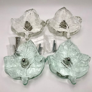 May include: Four clear glass leaf-shaped cabinet knobs with silver-toned hardware. The knobs have a textured surface and are accompanied by screws for installation. These decorative knobs add a natural touch to furniture.