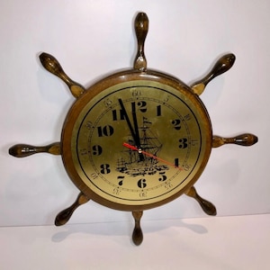 Puede incluir: Un reloj de rueda de barco de madera con una esfera dorada. La esfera del reloj presenta números negros, una ilustración de un barco y manecillas negras y rojas. El reloj tiene ocho radios de madera y está diseñado para parecerse a un timón de barco.