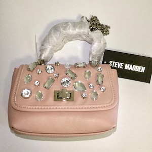 May include: A blush pink handbag with a top handle and a silver chain strap. The flap is adorned with various sizes of clear, sparkling rhinestones. A black tag with the text "STEVE MADDEN" is attached.