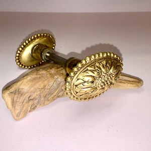 Könnte beinhalten: Ein Paar ovale Türknäufe aus Messing mit einem detaillierten geprägten Blumenmuster auf der Vorderseite und einem Perlenrand. Die beiden Knäufe sind durch einen zentralen Metallstift verbunden und haben ein goldenes Finish.