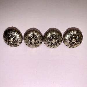 Peut inclure: Quatre boutons ronds argentés avec un motif symétrique détaillé. Chaque bouton présente un point central en relief entouré de lignes courbes et de formes en forme de pétales, créant une esthétique décorative et vintage.