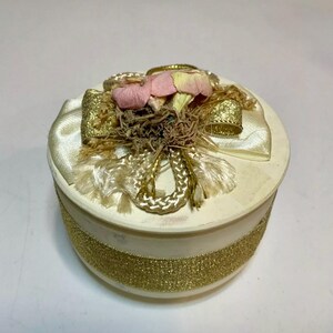 May include: A white round trinket box with a gold ribbon and a pink and white flower embellishment.