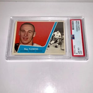 Könnte beinhalten: Eine 1963 Topps Reg Fleming Hockeykarte, bewertet mit EX-MT 6, in einer Schutzhülle. Die Karte zeigt ein Porträt von Fleming auf rotem Hintergrund und eine Actionaufnahme. Die Karte stammt von den Chicago Black Hawks.