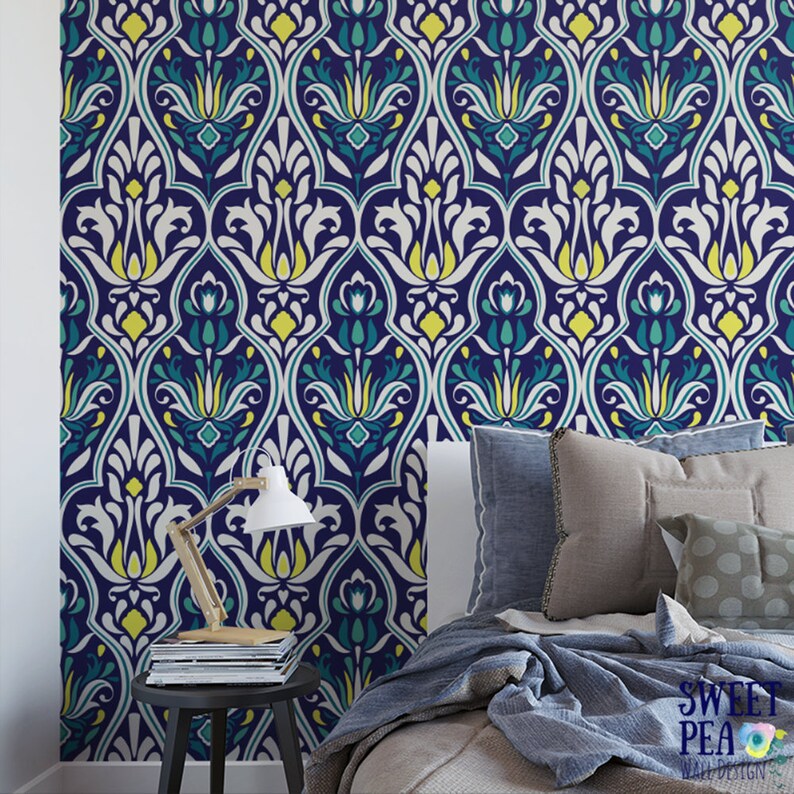 May include: A navy blue wallpaper with a repeating floral pattern in white, yellow, and teal. The wallpaper is installed on a wall behind a bed with pillows and a blanket. A small table with a lamp and magazines is in front of the wall.