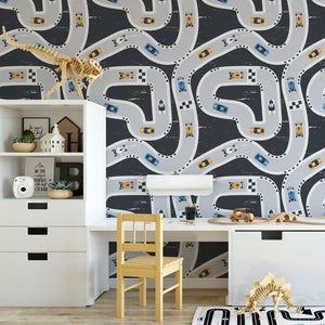 May include: A black and white race track wallpaper with yellow and blue cars. The wallpaper is in a child's room with a white desk and chair.
