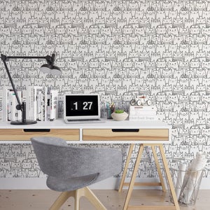 K&ouml;nnte beinhalten: Ein wei&szlig;er Schreibtisch mit einem Laptop, einer Lampe, B&uuml;chern und einer Pflanze. Die Wand hinter dem Schreibtisch ist mit einer schwarz-wei&szlig;en Tapete mit einem sich wiederholenden Muster von Katzen mit H&uuml;ten und Brillen bedeckt. Der Schreibtisch hat zwei Schubladen und einen Holzrahmen. Ein grauer B&uuml;rostuhl steht vor dem Schreibtisch. Der Schreibtisch steht auf einem Holzboden.