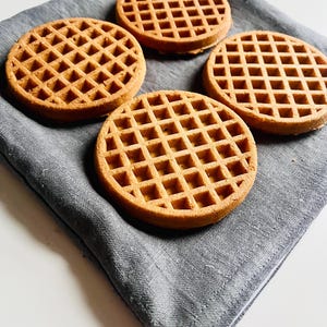 May include: Four round waffles with a grid pattern are arranged on a gray cloth. The waffles are golden brown and appear freshly made. The cloth is a muted gray color, providing a neutral backdrop for the waffles.