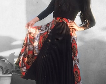 May include: A woman wearing a black long-sleeved shirt with a black pleated skirt with a red, brown, and white patterned overlay. She is wearing black heeled boots.
