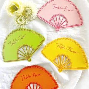 Peut inclure: Quatre marque-places en forme d'éventail de couleurs vives. Chaque carte est d'une couleur différente : rose, vert, jaune et orange. Chaque carte présente un motif d'éventail décoratif et les mots "Table One", "Table Two", "Table Three" et "Table Four".