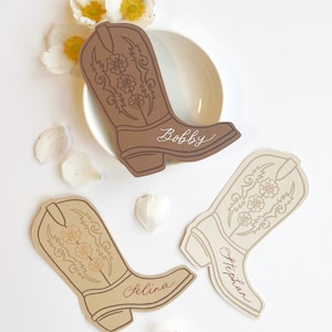 May include: Three cowboy boot-shaped place cards in shades of brown and beige, each with a name written in cursive. The boots feature floral designs and are arranged with white flowers and petals on a white surface.