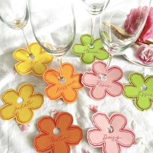 May include: A collection of colorful flower-shaped wine glass charms. Each charm is a different color, including yellow, orange, pink, and green, with a name written on each. The charms are arranged on a white surface, with wine glasses placed on top.