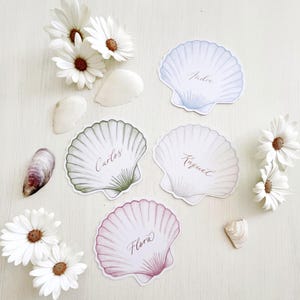 Tarjetas de mesa personalizadas con forma de concha de vieira: caligrafía dorada, decoración para bodas costeras.