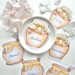 Puede incluir: Tarjetas de mesa con forma de tarro de miel con nombres escritos. Las tarjetas presentan ilustraciones de tarros de miel con miel goteando y abejas. Los nombres "Amelia", "Carlo", "Francesca", "Manaia", "Dominic" y "Daisy" están escritos en las tarjetas.
