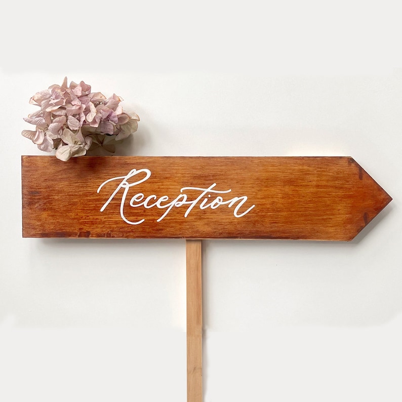 K&ouml;nnte beinhalten: Ein h&ouml;lzernes Pfeilschild mit dem Wort "Reception" in wei&szlig;er Kalligraphie. Das Schild ist mit einem Strau&szlig; blassrosa Blumen verziert.
