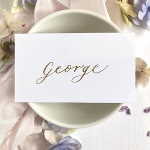 Könnte beinhalten: Eine weiße Tischkarte mit dem Namen "George" in goldener Kalligraphie.