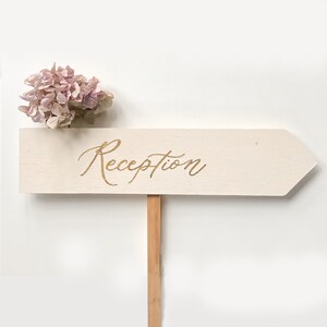 K&ouml;nnte beinhalten: Ein wei&szlig;es, h&ouml;lzernes Pfeilschild mit dem Wort "Reception" in goldener Kalligraphie. Das Schild ist mit einem Strau&szlig; getrockneter rosa Blumen verziert.