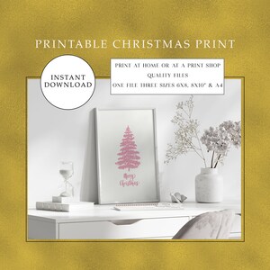 Könnte beinhalten: Druckbarer Weihnachtsdruck mit einer rosa Silhouette eines Weihnachtsbaums mit dem Text "Frohe Weihnachten" darunter. Der Druck ist in einem weißen Rahmen auf einem weißen Schreibtisch mit einer Vase mit weißen Blumen und einer weißen Sanduhr ausgestellt.