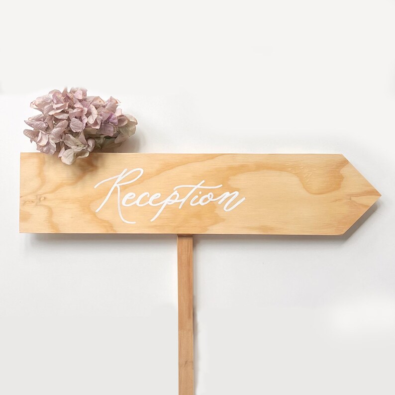 K&ouml;nnte beinhalten: Ein h&ouml;lzernes Pfeilschild mit dem Wort "Reception" in wei&szlig;er Kalligraphie. Ein B&uuml;schel getrockneter rosa Blumen liegt auf dem Schild.