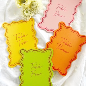 May include: Four colourful place cards with scalloped edges, labelled "Table One," "Table Two," "Table Three," and "Table Four." The cards are pink, yellow, orange, and green, with matching border colours. A small bouquet of white flowers sits nearby.