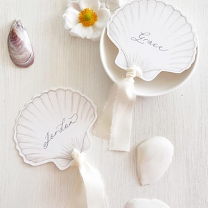 Puede incluir: Tarjetas de mesa en forma de concha con los nombres "Grace" y "Jordan" escritos en cursiva. Las tarjetas están atadas con cintas blancas, junto con flores blancas y conchas marinas. Un diseño delicado con temática playera.