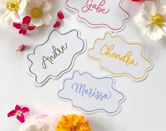 May include: White name cards with scalloped edges, each bearing a different name in cursive: Gabe, Andre, Chandra, and Marissa. The cards are surrounded by colorful flowers, including orange marigolds, white anemones, and red peonies, creating a vibrant, floral arrangement.