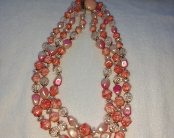 May include: A three-strand necklace with pink, peach, and clear beads. The necklace has a gold clasp.