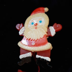 Può includere: Un ornamento di Natale vintage in plastica che rappresenta Babbo Natale con un abito rosso e bianco, barba bianca e occhi blu. L'ornamento è progettato per essere appeso a una corda.