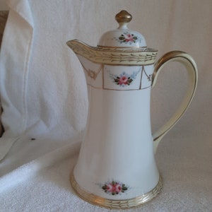 Puede incluir: Una cafetera de porcelana blanca con tapa, asa y boquilla. La cafetera está decorada con rosas rosas y ribetes dorados. La tapa tiene un adorno dorado. El asa y la boquilla son de color crema.