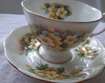 May include: A white teacup and saucer set with a floral pattern of yellow primroses and green leaves. The set has a gold trim around the edges.