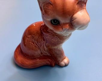 May include: A ceramic figurine of a seated orange tabby cat with green eyes. The cat has a white patch on its face and is posed with its head tilted downward. The figurine is on a light blue surface.