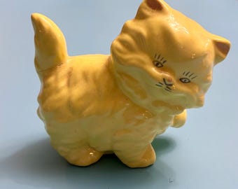 May include: A small, yellow ceramic kitten figurine with detailed facial features, including black painted eyes and whiskers. The kitten has a fluffy appearance and is posed in a walking stance. The figurine is likely a decorative item or collectible.