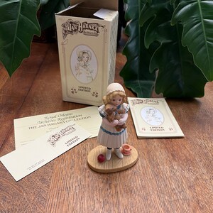 Könnte beinhalten: Eine Porzellanfigur eines jungen Mädchens in einem weißen Kleid, das einen Teddybär hält. Die Figur steht auf einem Holzsockel mit einem roten Apfel und einer Teetasse. Die Figur ist Teil der Jan Hagara Collection.