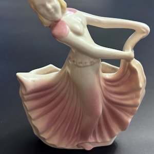May include: A ceramic figurine planter shaped like a dancer with blonde hair, wearing a cream-coloured bodice and a flowing, pleated skirt that transitions from cream at the waist to a soft pink hue at the hem, with pink accents on the shoulders.