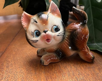 May include: A ceramic figurine of a brown, black, and white cat with blue eyes. The cat is sitting on its hind legs with its front paws outstretched. The cat has a playful expression on its face.