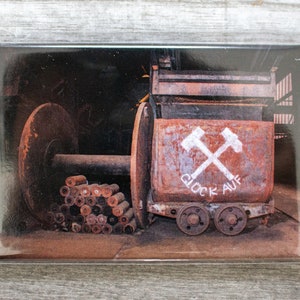Könnte beinhalten: Ein rostiger, alter Grubenwagen mit zwei gekreuzten Pickeln, die auf die Seite gemalt sind. Der Wagen trägt die Aufschrift "Glück Auf" in weißer Farbe. Der Wagen steht in einer dunklen, industriellen Umgebung.