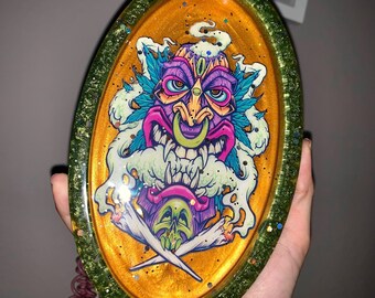 May include: A round, green and gold resin tray with a colorful illustration of a tiki-style face with a green, purple, and pink color scheme. The face is surrounded by green leaves and white smoke.