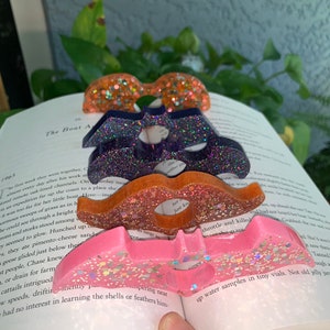 May include: A set of four colorful resin bookmark bookends shaped like mustaches. The mustaches are decorated with glitter and iridescent flakes. The mustaches are stacked on top of each other, with the pink mustache on the bottom and the orange mustache on the top.