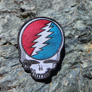 Könnte beinhalten: Gestickter Aufnäher mit einem stilisierten Totenkopf und Blitzmotiv. Das Design umfasst rote und blaue Bereiche mit einem weißen Blitz in der Mitte. Der Aufnäher hat einen schwarzen Rand und ist für Kleidung oder Accessoires geeignet.