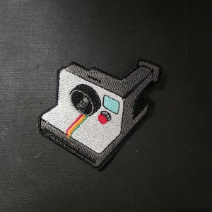 May include: Embroidered patch featuring a vintage instant camera design. The patch has a white body with a black lens and accents, a rainbow stripe, a red button, and a blue rectangle. The patch is a fun accessory for bags, jackets, or other items.