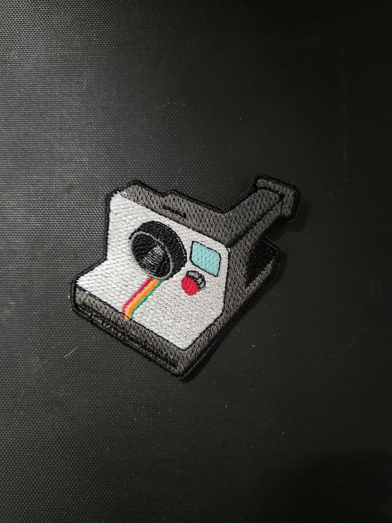 May include: Embroidered patch featuring a vintage instant camera design. The patch has a white body with a black lens and accents, a rainbow stripe, a red button, and a blue rectangle. The patch is a fun accessory for bags, jackets, or other items.