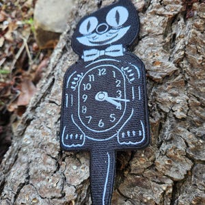 Puede incluir: Parche en blanco y negro con un gato de dibujos animados con pajarita, integrado en un diseño de reloj. La esfera del reloj muestra la hora y el parche tiene una cola larga y puntiaguda.