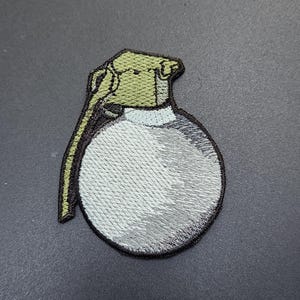 May include: Embroidered patch depicting a stylized grenade. The patch features a rounded body in shades of grey and white, with a green top and pull ring. The edges are outlined in black.