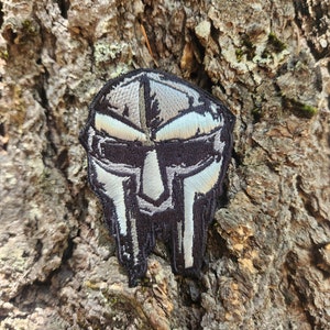 May include: Embroidered patch featuring a stylized mask design in black, silver, and white. The patch has a detailed, textured appearance and is likely intended for application on clothing or accessories. The patch is set against a textured brown background.